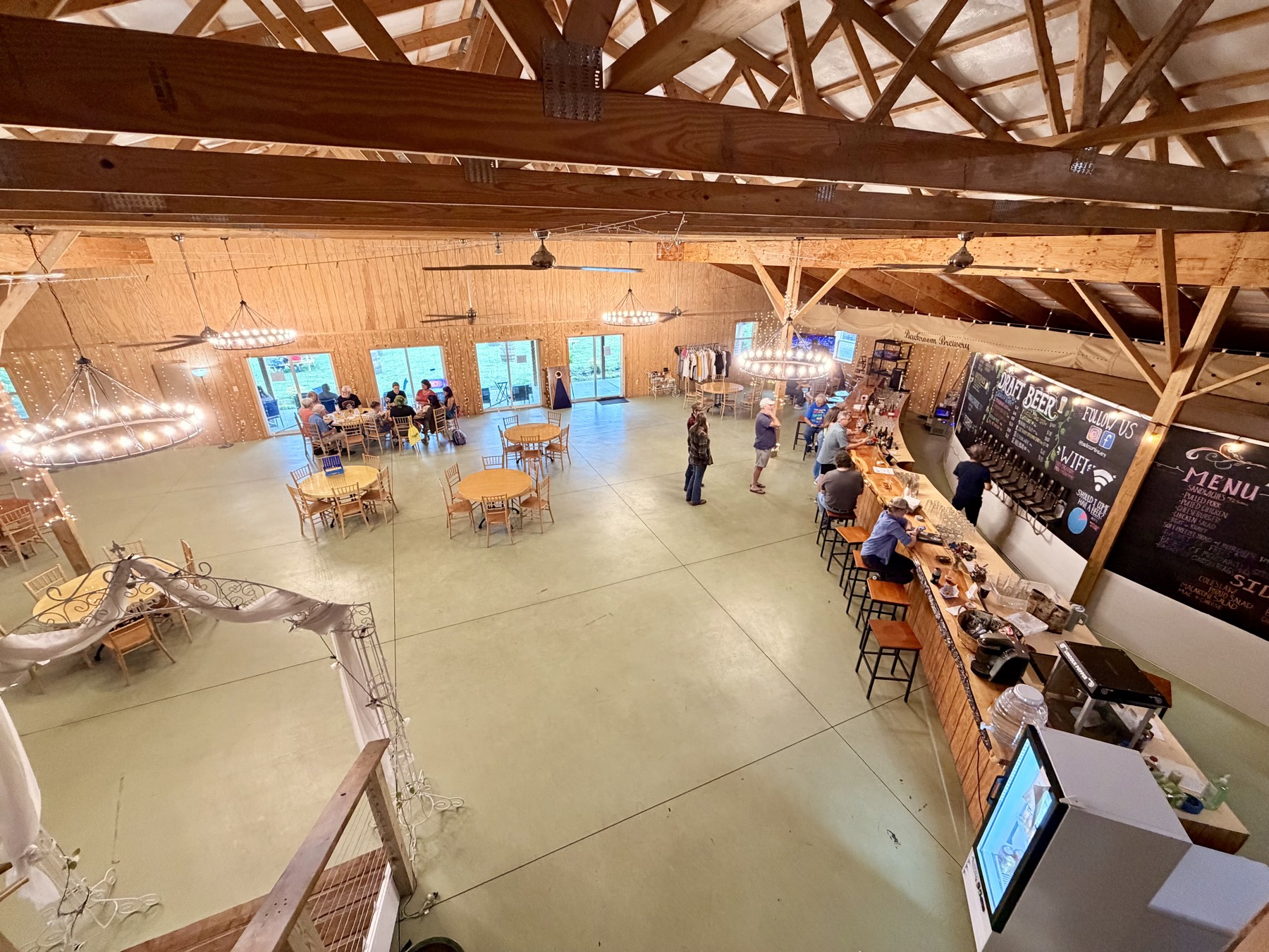 The Big Barn's main event space seen from the mezzanine — wagon-wheel chandeliers, round tables, and the live-edge bar